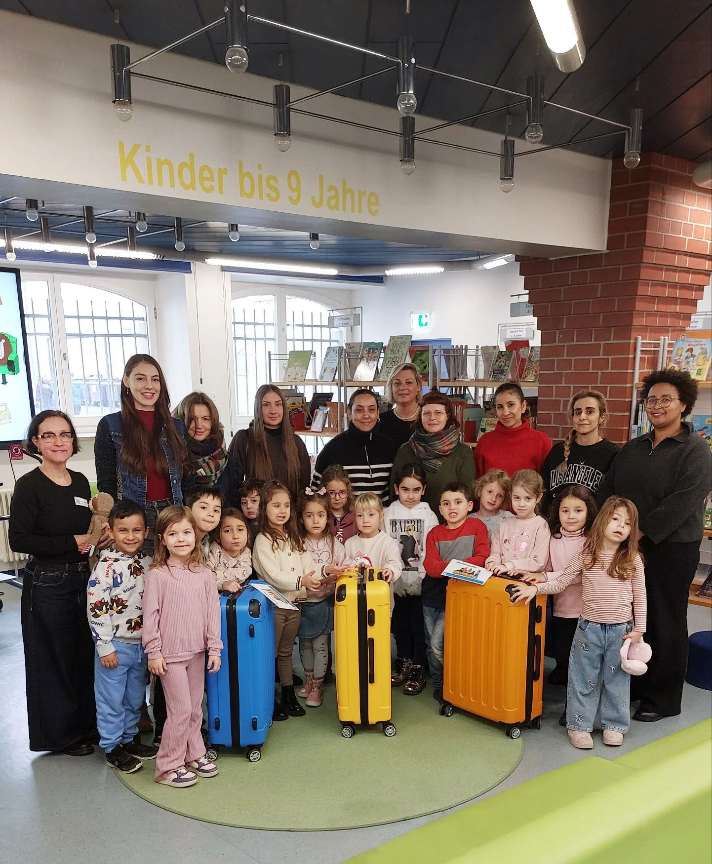 Das Bild zeigt eine Gruppe von 15 Kita-Kindern und zehn Erwachsenen bei der Übergabe von drei Medienkoffern in der Kinderbibliothek Offenbach. 